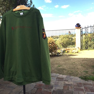 May include: A green long-sleeved sweatshirt with a red embroidered logo and a patch on the right sleeve. The sweatshirt is hanging on a hanger in front of a brick patio.