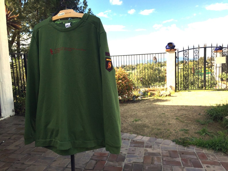 May include: A green long-sleeved sweatshirt with a red embroidered logo and a patch on the right sleeve. The sweatshirt is hanging on a hanger in front of a brick patio.