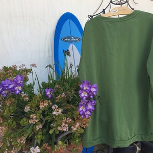 May include: A green long-sleeved sweatshirt hanging on a hanger in front of a white wall. A blue surfboard and a potted plant with purple flowers are in the background.