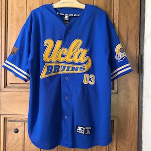 May include: Blue UCLA Bruins baseball jersey with yellow lettering and the number 03. The jersey has a button-up front, short sleeves, and a Starter logo. The sleeves have white and yellow stripes.