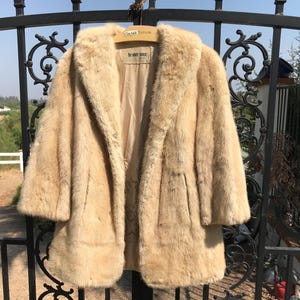 May include: A vintage, light brown faux fur coat with a wide collar and three-quarter sleeves. The coat is hanging on a black wrought iron gate. The coat has a satin lining and a label that reads "For the better dressed."