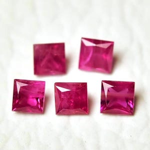 May include: Five square-cut, vibrant pink gemstones are arranged on a white surface. Each gem has a faceted surface, catching the light and displaying a rich, saturated color. The gems are likely rubies, perfect for jewelry making.