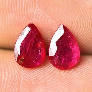 Könnte beinhalten: Zwei facettierte, tropfenförmige Rubine. Die Edelsteine sind ein tiefes, leuchtendes Rot mit einer polierten, reflektierenden Oberfläche. Die Rubine werden vor einem neutralen Hintergrund präsentiert, um ihre Farbe und ihren Schliff hervorzuheben.