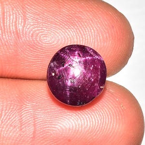May include: A round, polished ruby gemstone, exhibiting a deep magenta hue with internal star-like inclusions. The gem is held between two fingers, showcasing its size and luster. The gemstone is a focal point, highlighting its natural beauty.