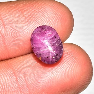 May include: An oval-shaped, cabochon-cut star ruby gemstone with a deep magenta-pink hue. The stone displays a distinct six-rayed star effect across its polished, translucent surface, featuring natural internal inclusions and dark speckles.