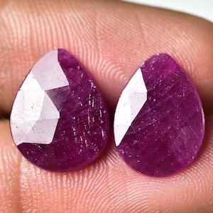 May include: Two faceted, teardrop-shaped gemstones with a deep magenta hue. The gems have a polished, reflective surface with multiple facets catching the light. The gems are held in a hand, showcasing their size and color.