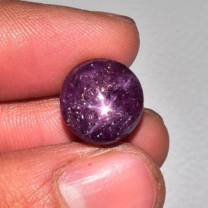 Op de afbeelding: Een ronde, gepolijste, dieppaarse edelsteen met een stervormig effect. De edelsteen heeft een glad oppervlak met een glinsterend, veelzijdig uiterlijk. Het ster-effect wordt gecreëerd door lichtreflectie van insluitsels in de steen.