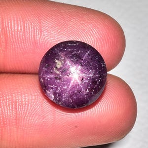 May include: A round, polished, purplish-red gemstone with a six-ray star effect. The gem is held between two fingers, showcasing its colour and the star-like pattern across its surface. The stone has a smooth, reflective surface.