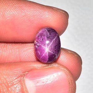 Peut inclure: Pierre précieuse en forme ovale, un rubis étoilé. La gemme est d'un magenta profond avec un motif en étoile à six rayons sur sa surface. La pierre est tenue entre les doigts, mettant en valeur ses qualités polies et réfléchissantes. La couleur du rubis varie du magenta clair au foncé.