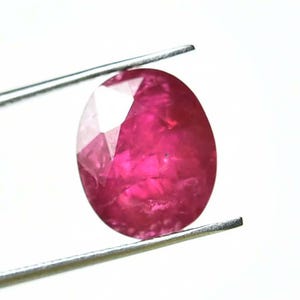 May include: A faceted, oval-shaped ruby gemstone, held by tweezers against a white background. The ruby displays a deep, vibrant red color with internal light reflections. The tweezers are silver.