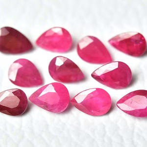 May include: A collection of ten faceted, teardrop-shaped rubies. The gemstones display a range of deep red to vibrant pink hues, with multiple facets catching the light. The rubies are arranged on a white surface.