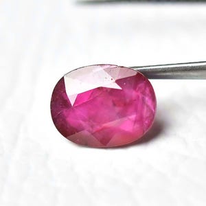 May include: An oval-shaped, faceted ruby gemstone. The ruby is a deep pinkish-red color, with a polished surface that reflects light. The gemstone is held by a pair of metal tweezers against a white background.