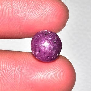 May include: A close-up of a round, polished gemstone. The gem is a deep magenta color with small black inclusions. The stone is held between two fingers, showcasing its size and shape. The gem appears to be a ruby.