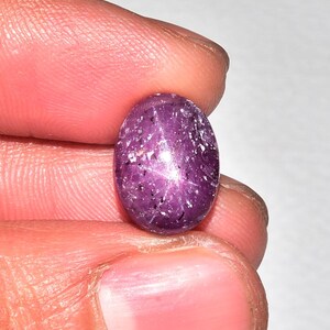 Può includere: Gemma ovale rosso-violacea con effetto a stella. La superficie lucida della gemma presenta piccole inclusioni. La pietra è tenuta tra le dita. Il colore è una tonalità profonda e ricca, e l'effetto a stella è visibile.