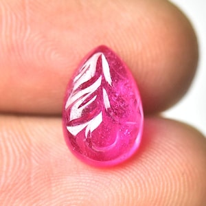 May include: A teardrop-shaped, polished, fuchsia-colored gemstone with a carved white leaf design. The gem is held between two fingers, showcasing its vibrant color and intricate detailing. The gem is a cabochon.
