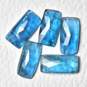 May include: Five faceted, rectangular blue gemstones with a polished finish. The gems are a vibrant, light blue colour and have a geometric cut. The gemstones are arranged on a white surface.
