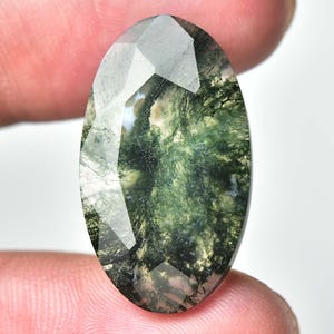 May include: Oval-shaped moss agate gemstone with a faceted surface. The gem features a translucent appearance with green, white, and brown inclusions, resembling a miniature landscape. Held between fingers against a white background.