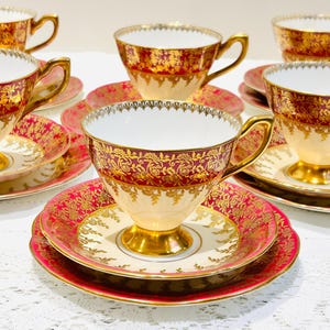 May include: A set of vintage teacups and saucers. The teacups have a cream-colored base with red and gold floral patterns. The saucers and plates are red with gold trim. The teacups have gold handles and footed bases.