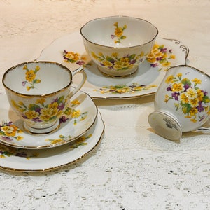 May include: A vintage tea set featuring a teacup, saucer, and a small plate, all adorned with a floral pattern in yellow and purple. The edges of the set are trimmed with gold. The set is placed on a white lace tablecloth.