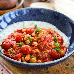 Puede incluir: Un cuenco azul moteado lleno de una base cremosa blanca, cubierto con un guiso vibrante de tomate y frijoles. El perejil fresco adorna el plato. La comida se presenta sobre una superficie de madera.
