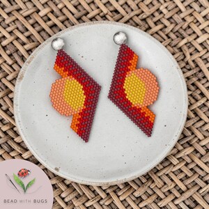 Puede incluir: Un par de pendientes geométricos hechos de pequeñas cuentas en tonos rojo, naranja y amarillo. Cada pendiente presenta un poste de color plateado y una forma angular puntiaguda con una sección circular integrada en el diseño.