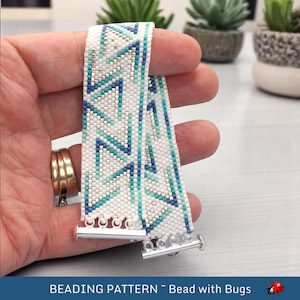 May include: A white beaded bracelet with a geometric pattern in shades of blue and silver. The bracelet has silver toggle clasps.