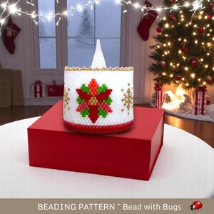 May include: A white beaded candle holder with a red poinsettia design and gold accents. The candle holder is sitting on a red box and a white surface. The background includes a Christmas tree and a window with lights.