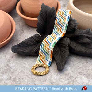 May include: A white beaded bracelet with a geometric pattern in shades of blue, green, gold, and orange. The bracelet has a gold metal ring clasp. The bracelet is lying on a black leaf.