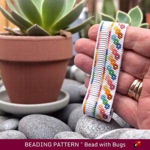 Può includere: Un braccialetto bianco di perline con un motivo colorato di piccole perline. Il motivo presenta una fila di piccoli cerchi colorati e una fila di cerchi colorati più grandi. Il braccialetto è tenuto in una mano.