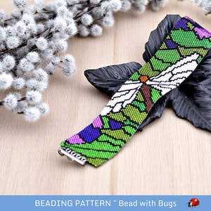 Puede incluir: Un brazalete de cuentas con un diseño de libélula colorido. El brazalete es verde, blanco, morado y naranja. La libélula es blanca con alas marrones. El brazalete tiene un cierre plateado.