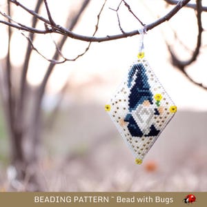 以下が含まれることがあります： 青い帽子と黄色の花を持つノームのデザインの、ビーズで飾られたひし形のオーナメント。オーナメントは白で、金と黄色のアクセントがあり、木の枝から吊り下げられています。下部のテキストは「Beading Pattern ~ Bead with Bugs」と表示されています。
