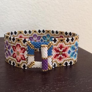 May include: A colorful beaded bracelet with a geometric pattern and a gold clasp. The bracelet is made with small, colorful beads in a variety of shades, including pink, blue, red, and white. The beads are arranged in a geometric pattern, creating a visually appealing design. The bracelet has a gold clasp that is secured with a small, metal loop.
