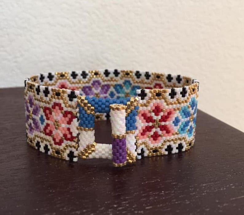 May include: A colorful beaded bracelet with a geometric pattern and a gold clasp. The bracelet is made with small, colorful beads in a variety of shades, including pink, blue, red, and white. The beads are arranged in a geometric pattern, creating a visually appealing design. The bracelet has a gold clasp that is secured with a small, metal loop.