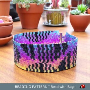 May include: A beaded bracelet with a colorful, abstract pattern in shades of pink, purple, blue, and black. The bracelet is made with small, seed beads and has a metal clasp.