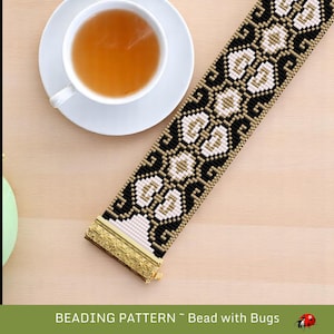 May include: A beaded bracelet featuring a pattern of white, black, and gold beads. The bracelet has a gold clasp and is next to a cup of tea on a saucer. The text "BEADING PATTERN ~ Bead with Bugs" is at the bottom.