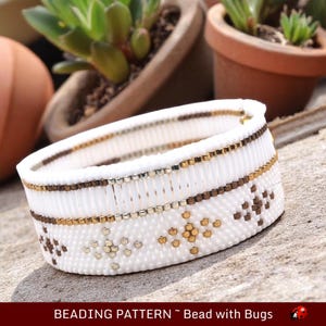 Op de afbeelding: Witte kralen armband met een geometrisch patroon van bruine, gouden en zilveren kralen. De armband ligt op een houten oppervlak. "Beading Pattern - Bead with Bugs" staat onder het armband.