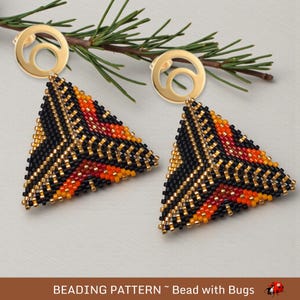 May include: A pair of gold-colored earrings with a circular top and a triangular beaded pendant. The pendant features a black, orange, red, and gold pattern. The earrings are displayed on a light-colored surface with a sprig of greenery.