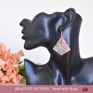 May include: Beaded earrings in the shape of a triangle with a floral pattern in pink, orange, yellow, and green. The earrings are hanging from a mannequin's ear.