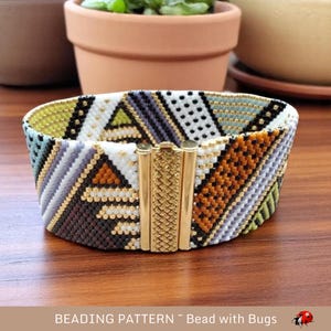 May include: A beaded bracelet with a geometric pattern in shades of white, black, orange, green, and gold. The bracelet has a gold clasp.