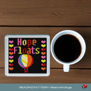 Puede incluir: Un posavasos cuadrado con cuentas con las palabras "Hope Floats" y un diseño de globo aerostático colorido. El posavasos está junto a una taza de café blanca llena de café negro, todo sobre una superficie de madera.