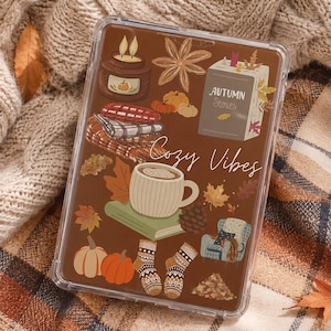 Könnte beinhalten: Eine transparente Tablethülle mit braunem Hintergrund und herbstlichen Illustrationen wie einer Kerze, einem Buch mit dem Titel Autumn Stories, einer Kaffeetasse, Kürbissen, gemusterten Socken, einem Sessel und dem Text Cozy Vibes in Weiß.