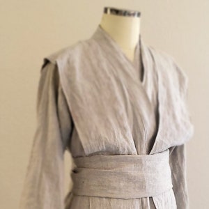 May include: A light gray linen robe with a long sash tied at the waist. The robe has a V-neck and long sleeves.