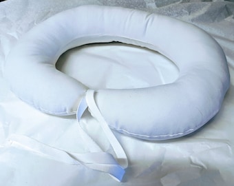 May include: White donut-shaped pillow with a white fabric strap. The pillow is made of soft, plush fabric and is designed to provide support and comfort.