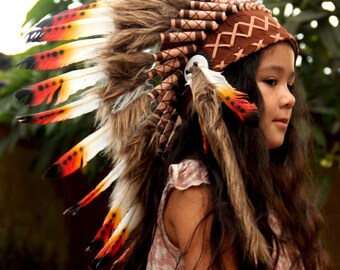 Puede incluir: Un niño con un tocado de plumas marrones y blancas con detalles rojos y naranjas. El tocado tiene una banda de cuero marrón con un patrón tejido. El niño lleva un vestido floral rosa y blanco.