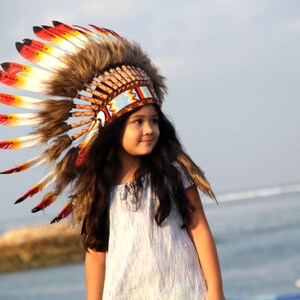 Puede incluir: Una ni&ntilde;a peque&ntilde;a con un tocado tradicional nativo americano con plumas rojas, amarillas y blancas. El tocado est&aacute; hecho de piel marr&oacute;n y tiene una banda de cuentas. La ni&ntilde;a lleva un vestido blanco y tiene el pelo largo y casta&ntilde;o. Est&aacute; de pie en una playa con el oc&eacute;ano de fondo.