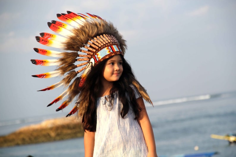 Puede incluir: Una ni&ntilde;a peque&ntilde;a con un tocado tradicional nativo americano con plumas rojas, amarillas y blancas. El tocado est&aacute; hecho de piel marr&oacute;n y tiene una banda de cuentas. La ni&ntilde;a lleva un vestido blanco y tiene el pelo largo y casta&ntilde;o. Est&aacute; de pie en una playa con el oc&eacute;ano de fondo.