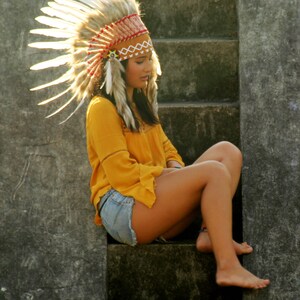 May include: A woman wearing a white and brown feathered headdress sits on concrete steps. She is wearing a yellow shirt and denim shorts.