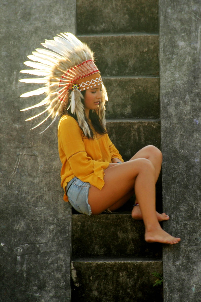 May include: A woman wearing a white and brown feathered headdress sits on concrete steps. She is wearing a yellow shirt and denim shorts.