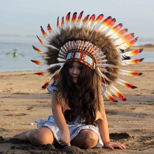 Puede incluir: Una persona joven con un tocado nativo americano colorido con plumas. El tocado est&aacute; hecho de plumas marrones, rojas y blancas. La persona lleva un vestido azul claro y tiene el pelo largo y casta&ntilde;o.