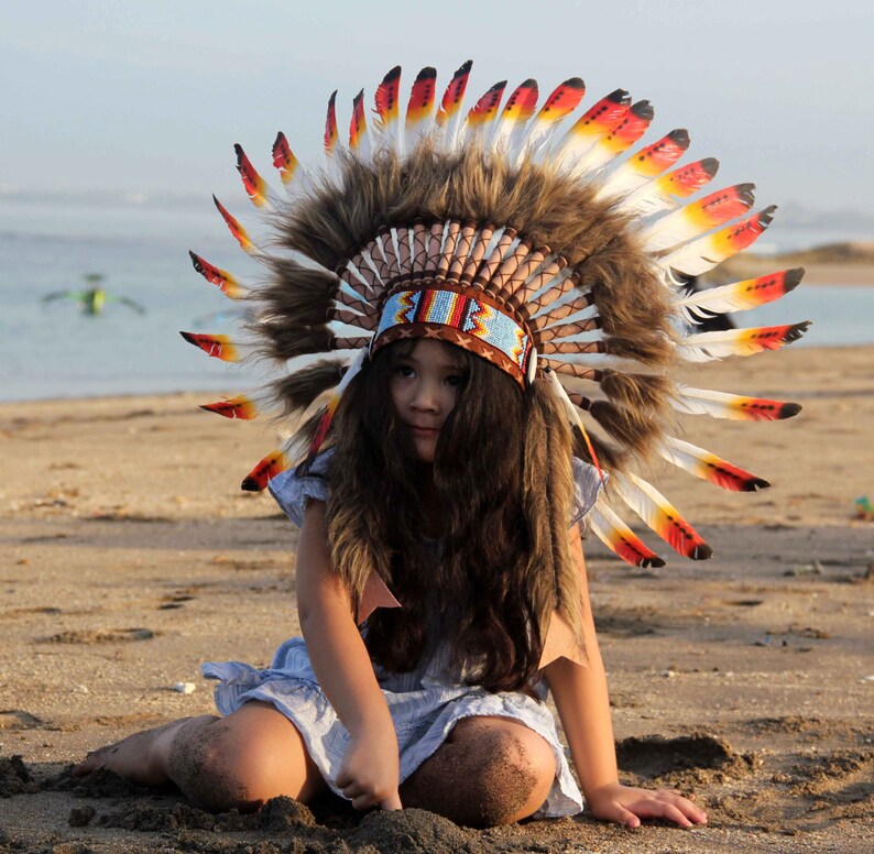Puede incluir: Una persona joven con un tocado nativo americano colorido con plumas. El tocado est&aacute; hecho de plumas marrones, rojas y blancas. La persona lleva un vestido azul claro y tiene el pelo largo y casta&ntilde;o.